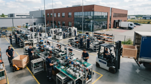Obraz przedstawia nowoczesną, przestronną halę produkcyjną połączoną z przestrzenią magazynową i strefą załadunku, zlokalizowaną w nowoczesnym budynku z cegły i szkła